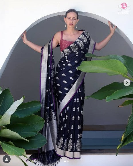 Woman wearing a navy blue saree with white patterns, standing against a neutral background with green plants.