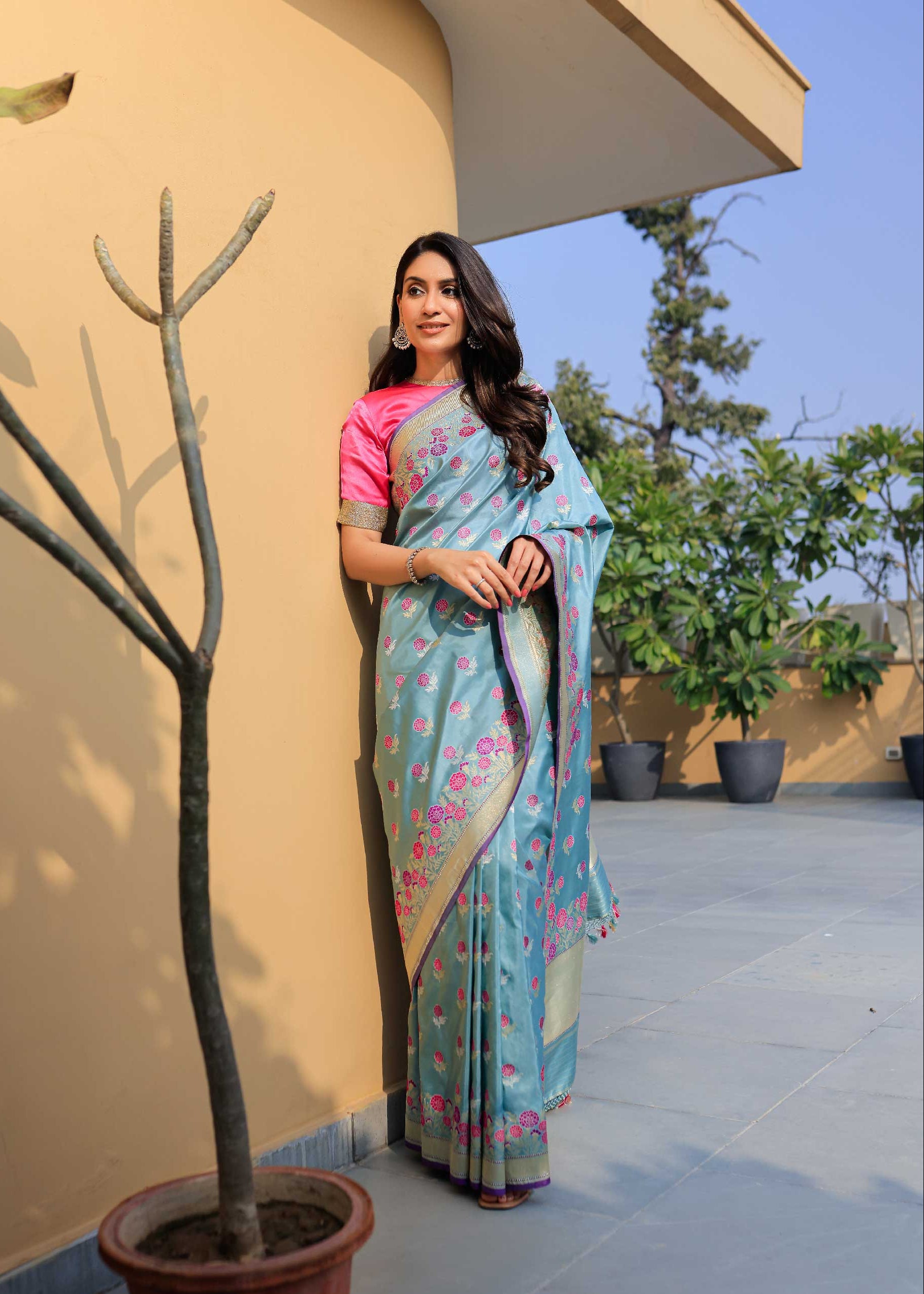 Woman in a blue saree with a pink blouse standing outdoors against a beige wall.
